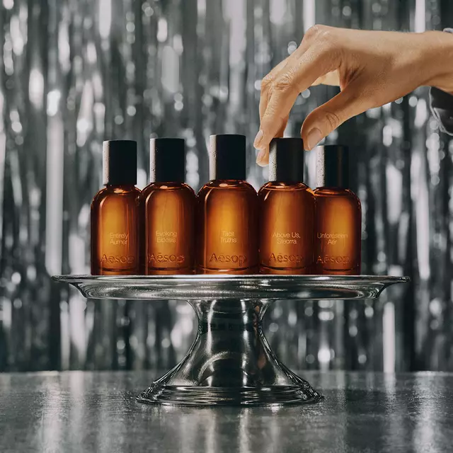 Amber Aesop fragrance bottles with black caps arranged among wooden blocks of different heights against a dark brown background.