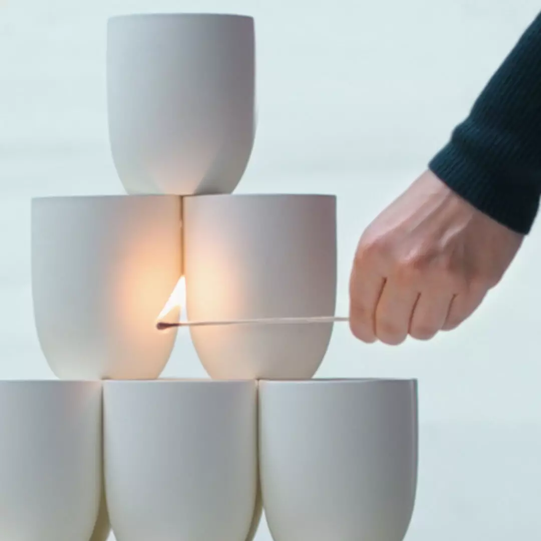 A hand holds a long burning match beside stacked white vessels, with a red fringed scarf at the right.