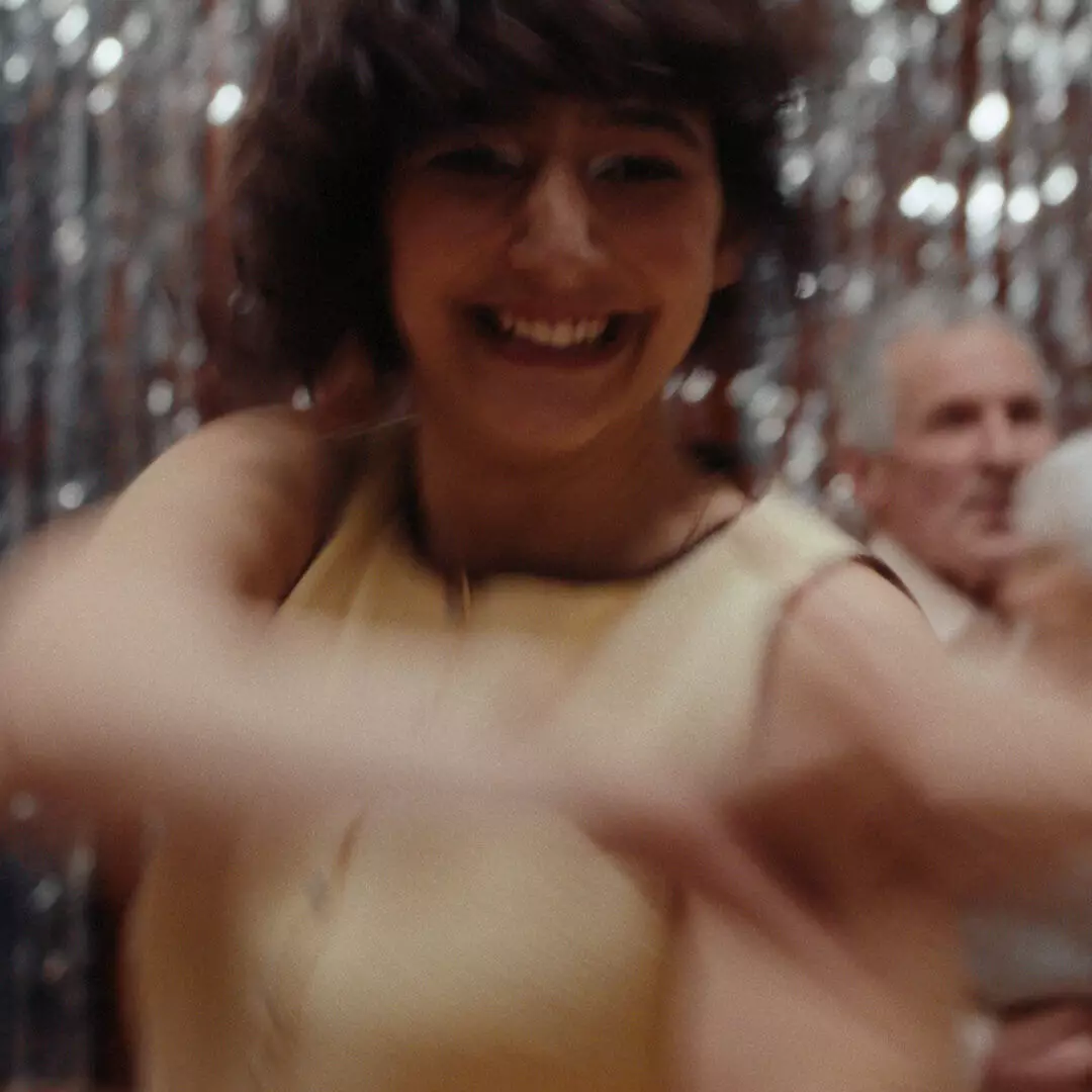 A smiling person in a pale yellow sleeveless top holds another person's hand, with people and silver streamers behind.