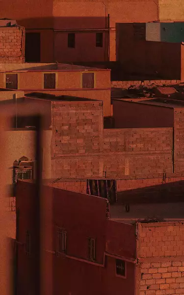 Terracotta city rooftops and walls in warm rust-red light, with blocky architecture, shadowed facades, and textured masonry surfaces.