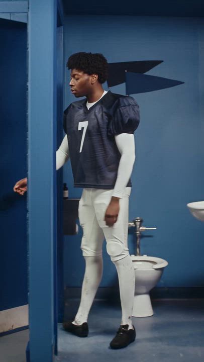 Football player in a navy jersey standing in a blue public restroom near a toilet