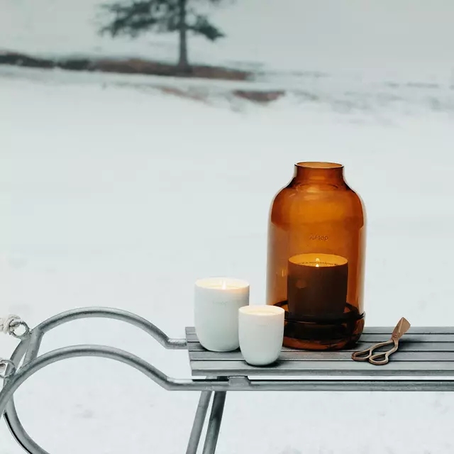Amber bottles, a white candle and brass-coloured vessels stand on a reflective surface before rocky hills, with incense smoke rising.