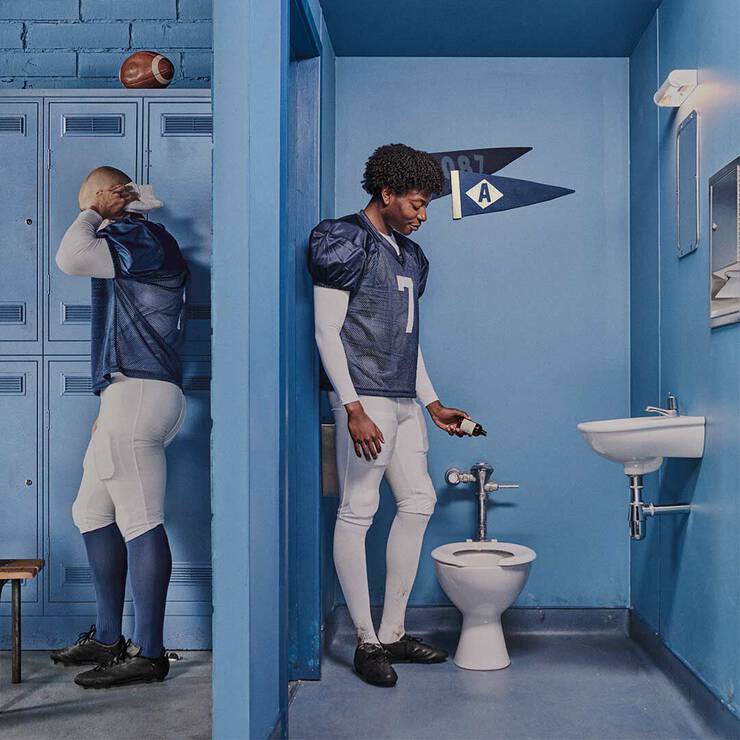 American football players in a blue locker room, one standing beside a toilet with Aesop Post-Poo Drops.