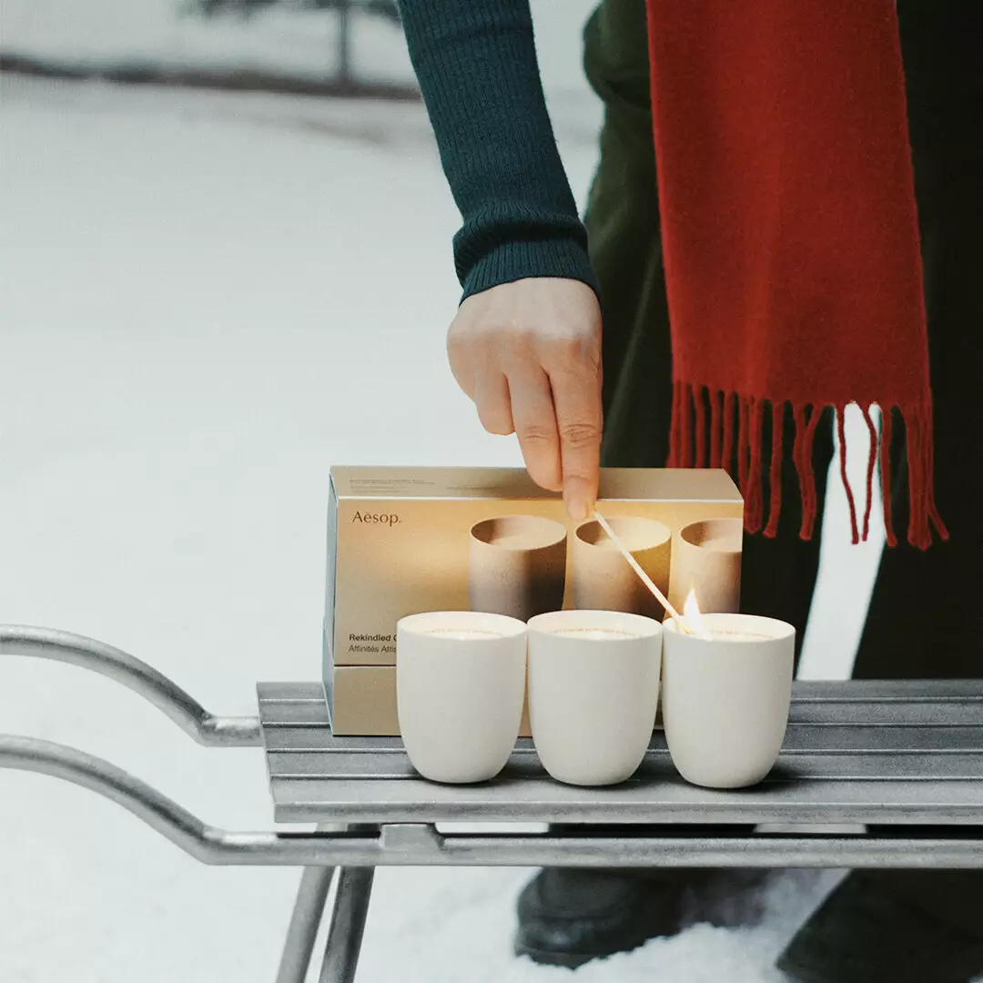 A person wearing a red scarf lights one of three white candles beside an Aesop box on a metal sledge in the snow.