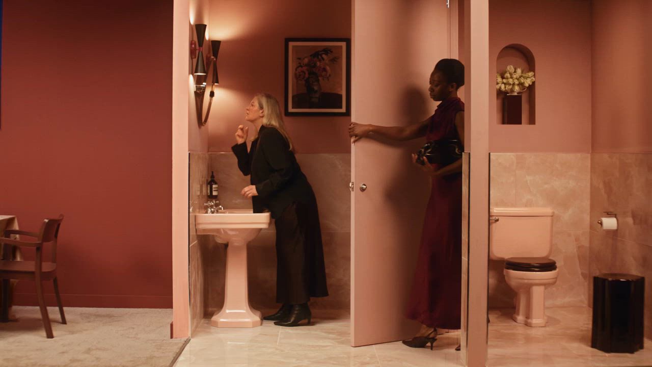 Woman in a burgundy dress entering a restroom stall while another woman stands at the sink