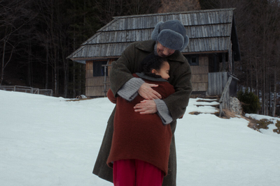 An adult in a dark coat and grey hat embraces a child in a rust-coloured coat outside a wooden cabin in the snow.