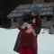 An adult in a dark coat and grey hat embraces a child in a rust-coloured coat outside a wooden cabin in the snow.