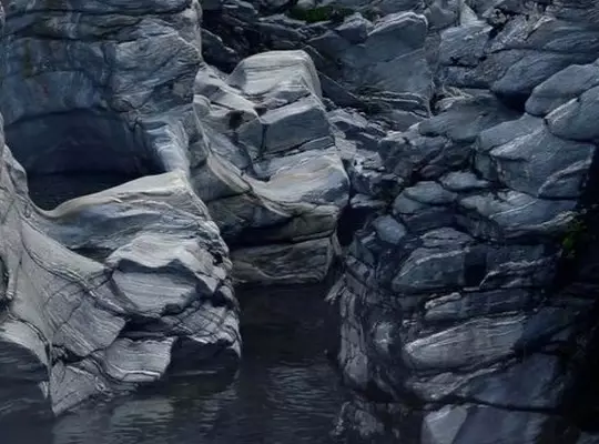 Layered slate-grey rock formations above dark water, showing striated mineral textures, cracks, and naturally sculpted contours.