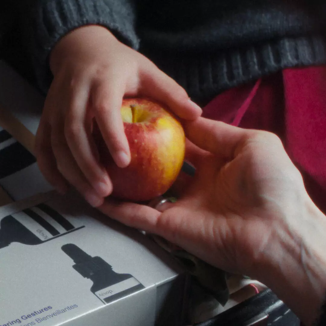 Two hands hold a red-and-yellow apple above Aesop boxes printed with black bottle and tube silhouettes, beside red fabric.