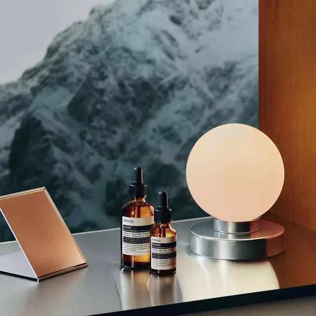 Amber Aesop bottles, jars and a silver tube are grouped beside a dark sink beneath an exposed metal tap, against a golden curtain.