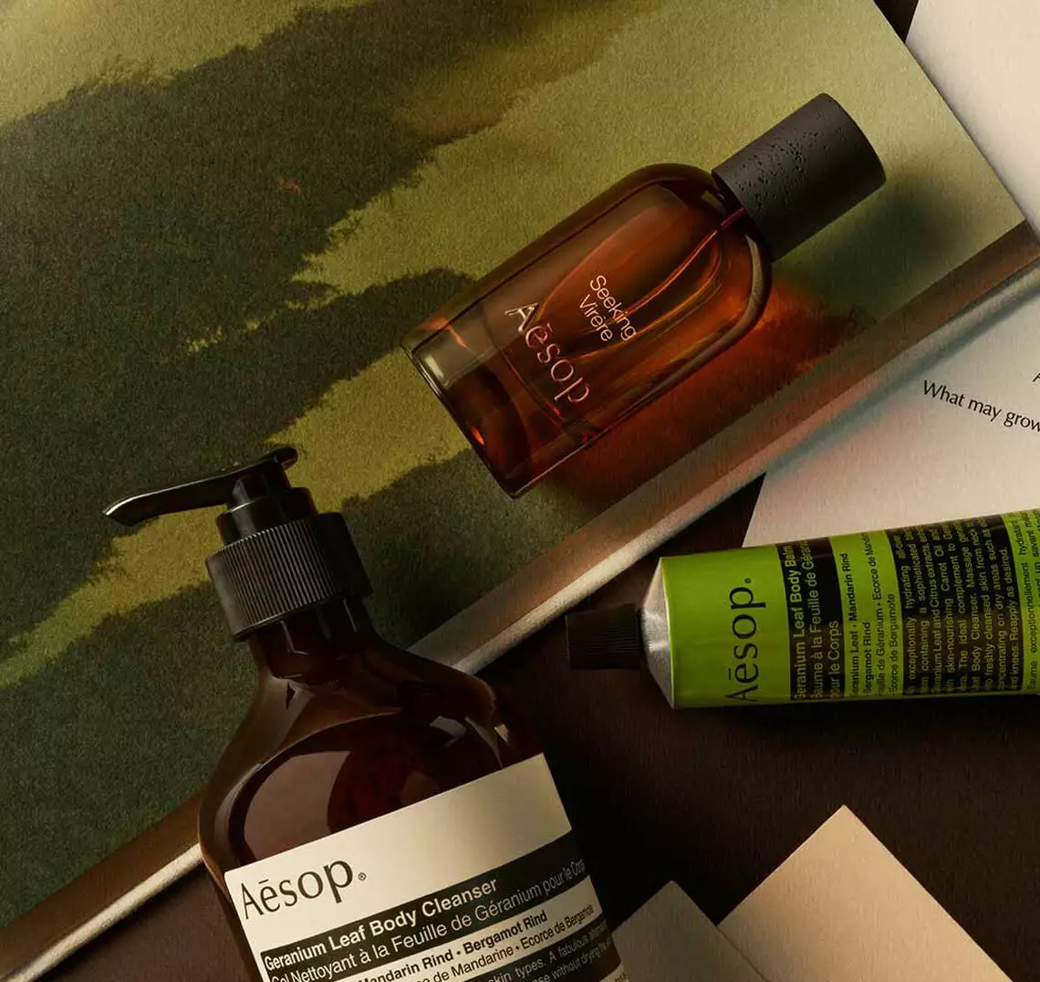 Aesop Seeking, Virere fragrance bottle, large pump bottle and cosmetic tube on an open book on a green textured background.