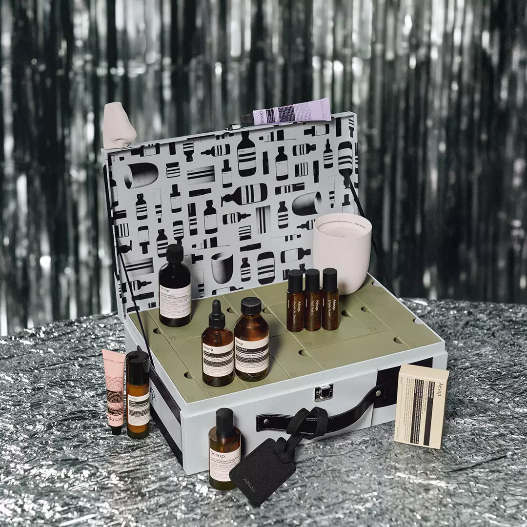 Aesop bottles, tubes and a white candle surround an open silver case with numbered green compartments and a patterned lid against silver tinsel.