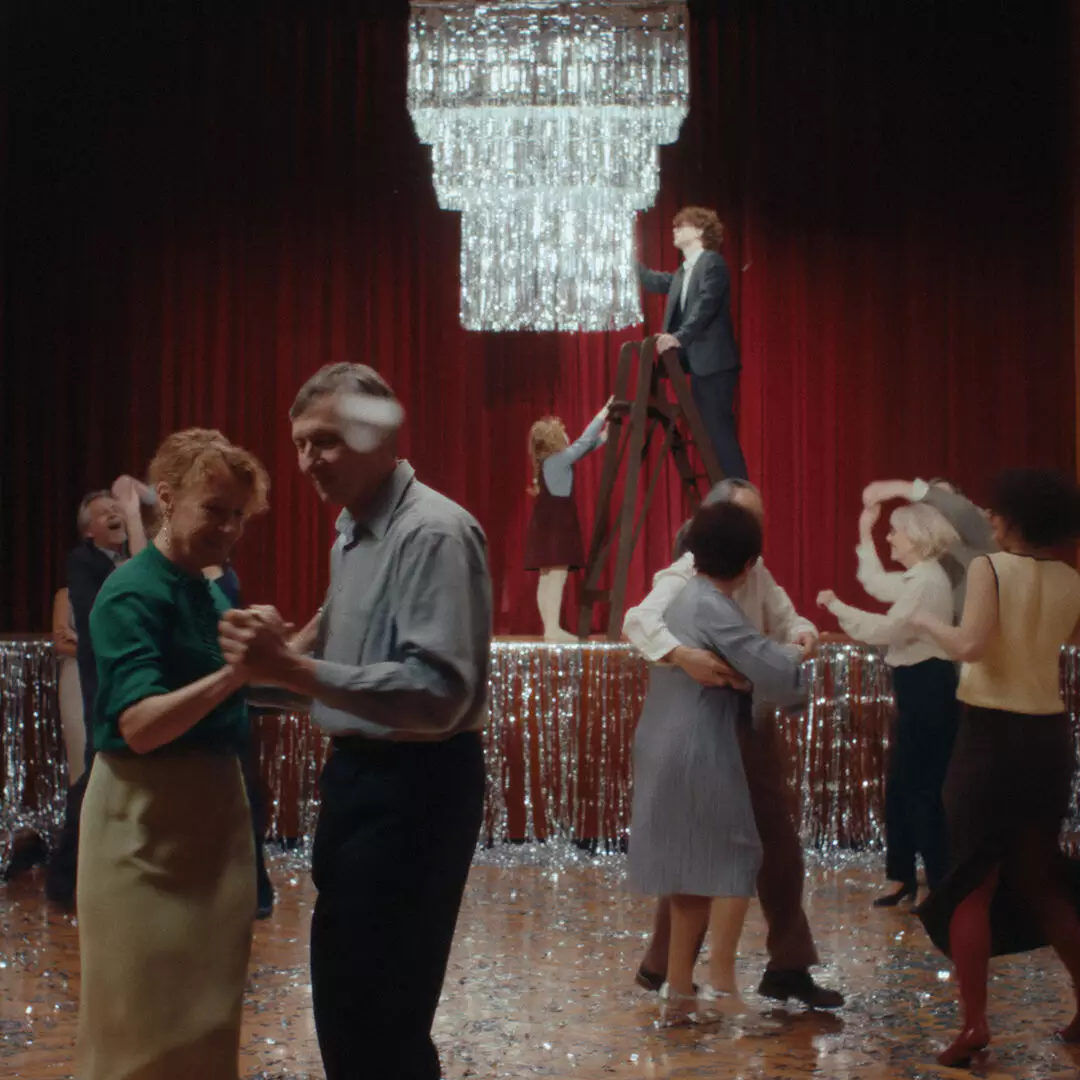 People dance beneath a large chandelier in a hall decorated with silver tinsel and red curtains, with a person standing on a ladder.