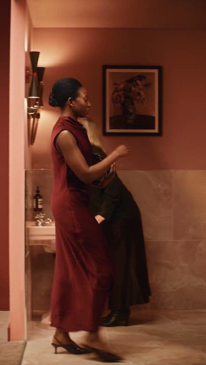 Woman in a burgundy dress standing in a warmly lit restroom beside a sink and wall art