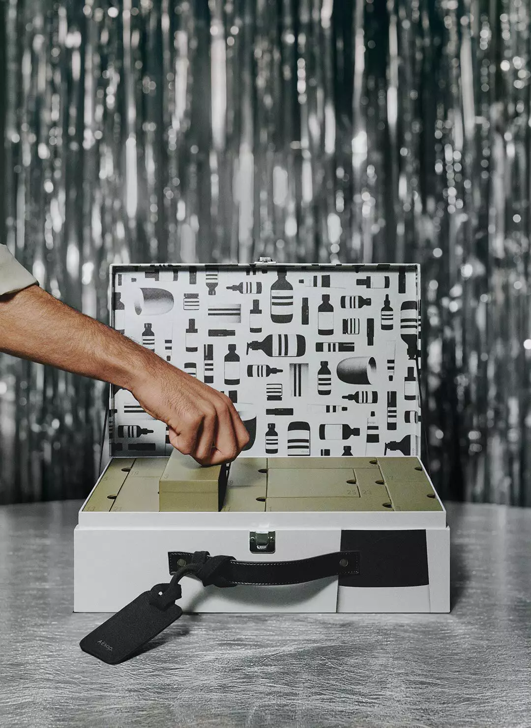 Hand lifting a green compartment lid inside an open silver case with a black-and-white patterned interior, on a table against silver tinsel.