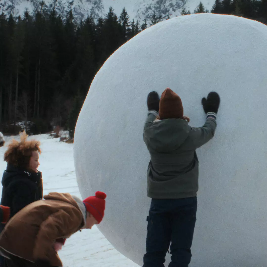 Four people in winter clothing gather beside a giant snowball, one pressing both gloved hands against it, with forest and mountains behind.