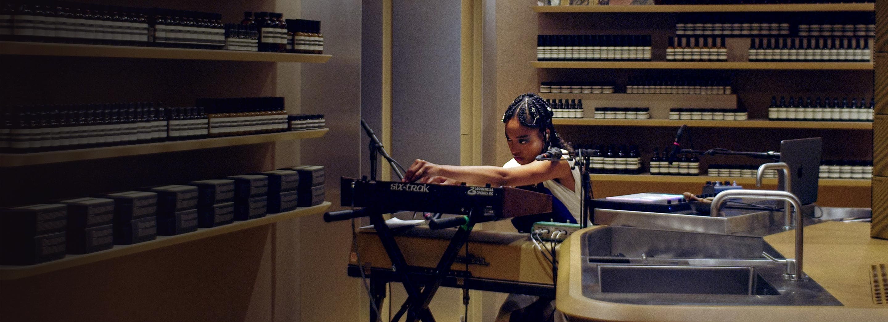 Kiala Ogawa operating a synthesizer surrounded by amber glass bottles in an Aesop store.