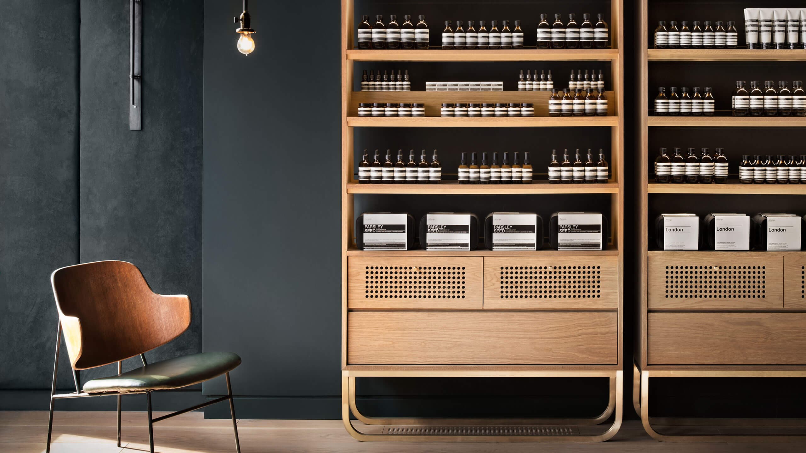 A mid-century chair sits underneath soft lighting adjacent to timber shelves holding Aesop Product range.