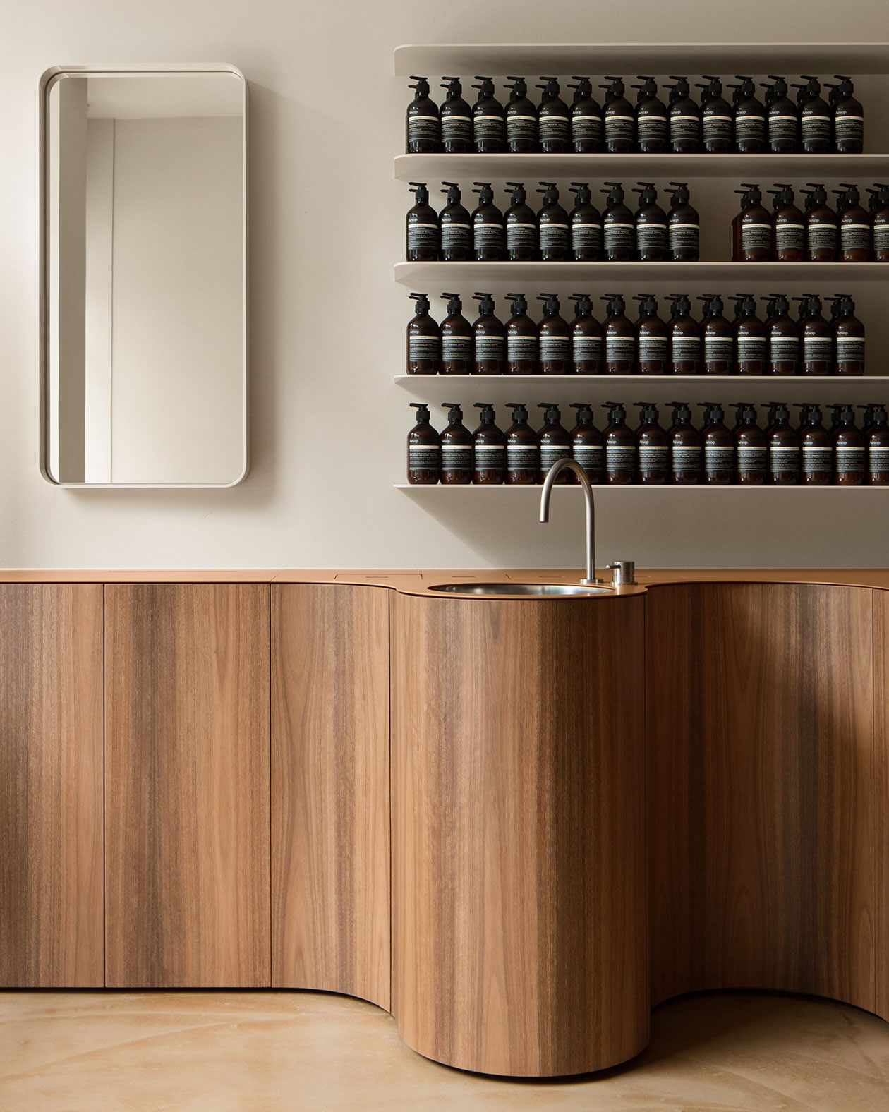 A wooden bench that has a basin installed. Several rows of shelving are hung on the wall behind displaying Aesop products.