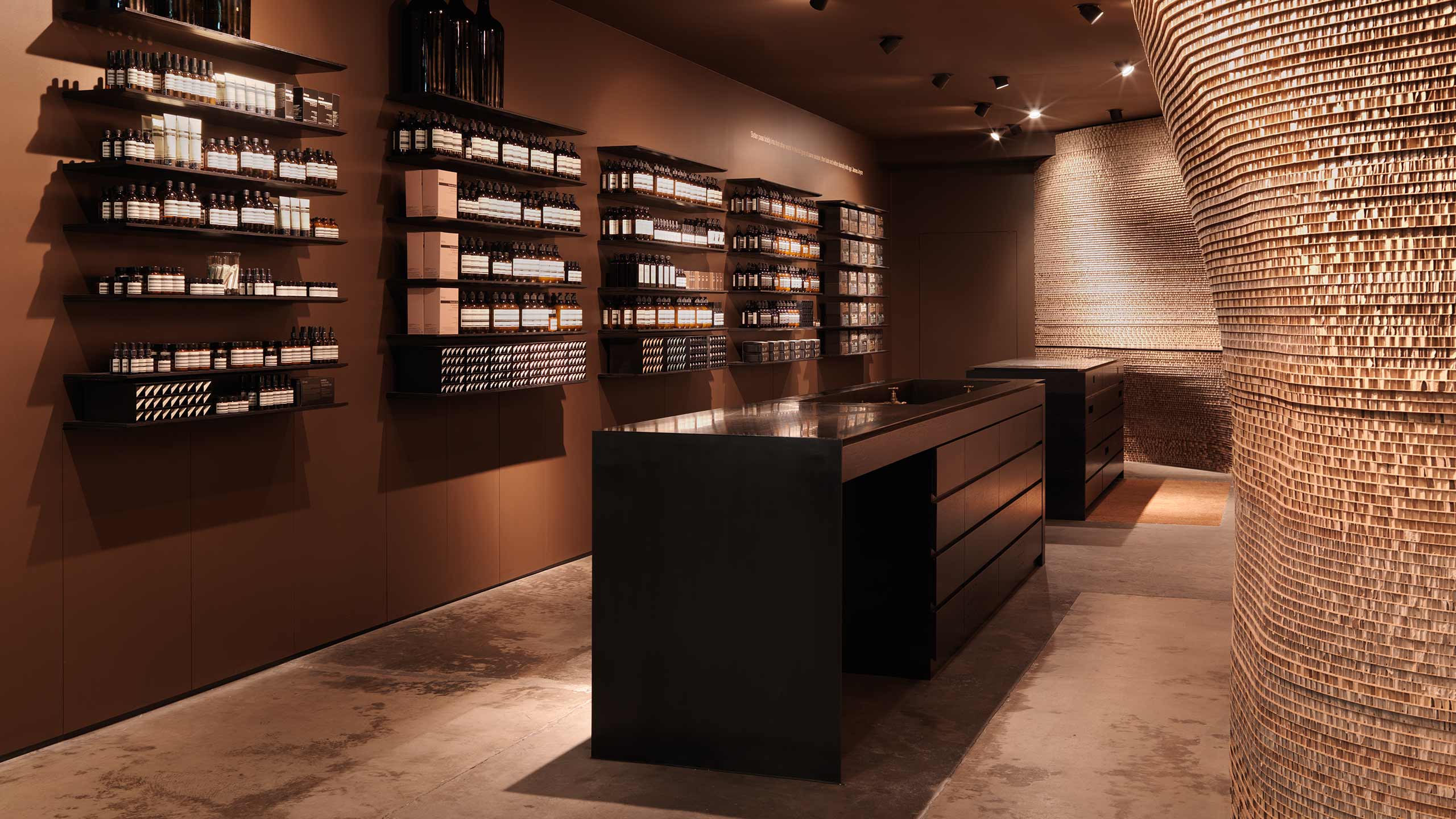 A dark, brown, brassy wall with floating shelving that displays a vast array of different Aesop products.