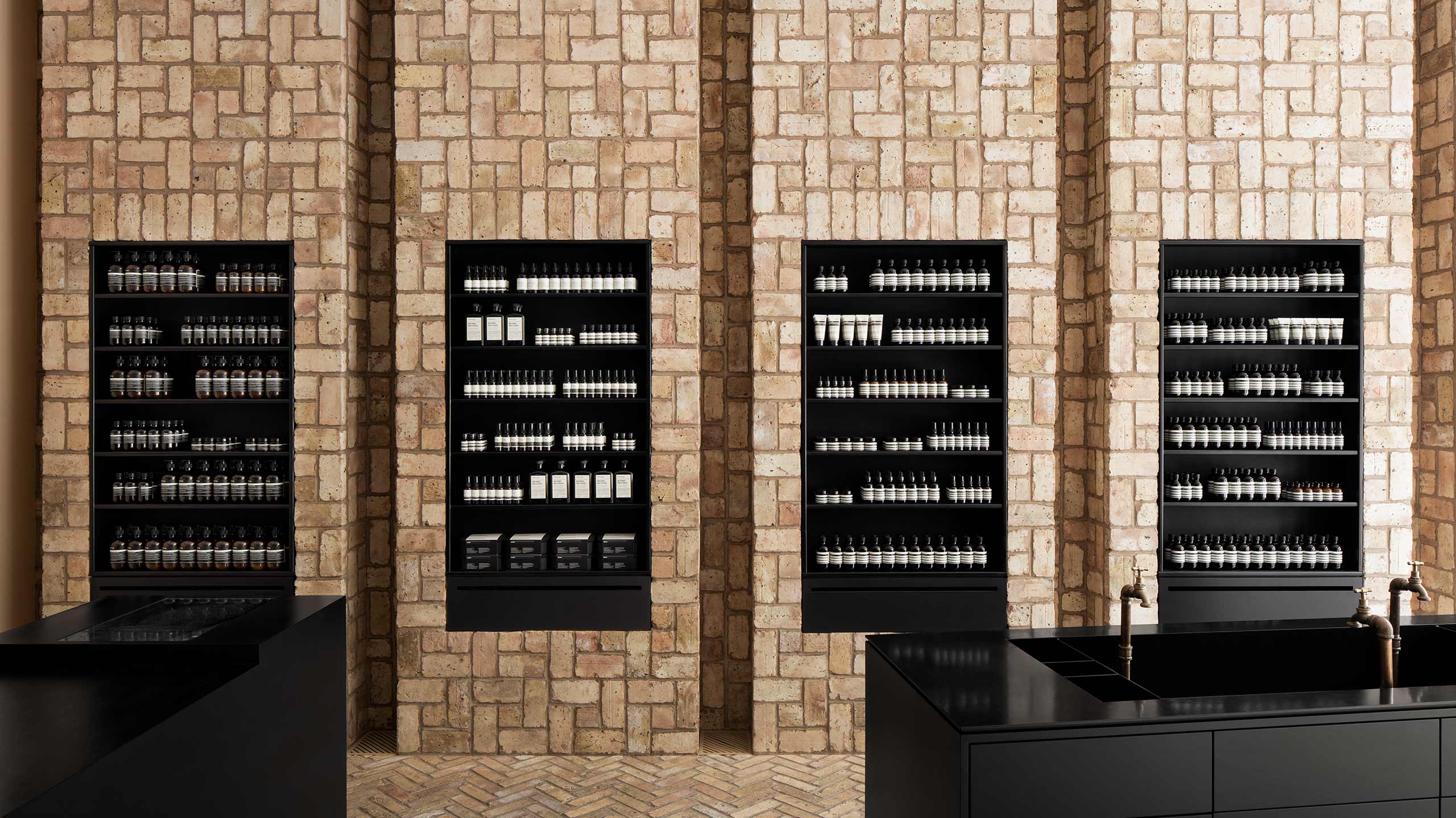 Four brick walls fitted with black steel shelves holding Aesop product.