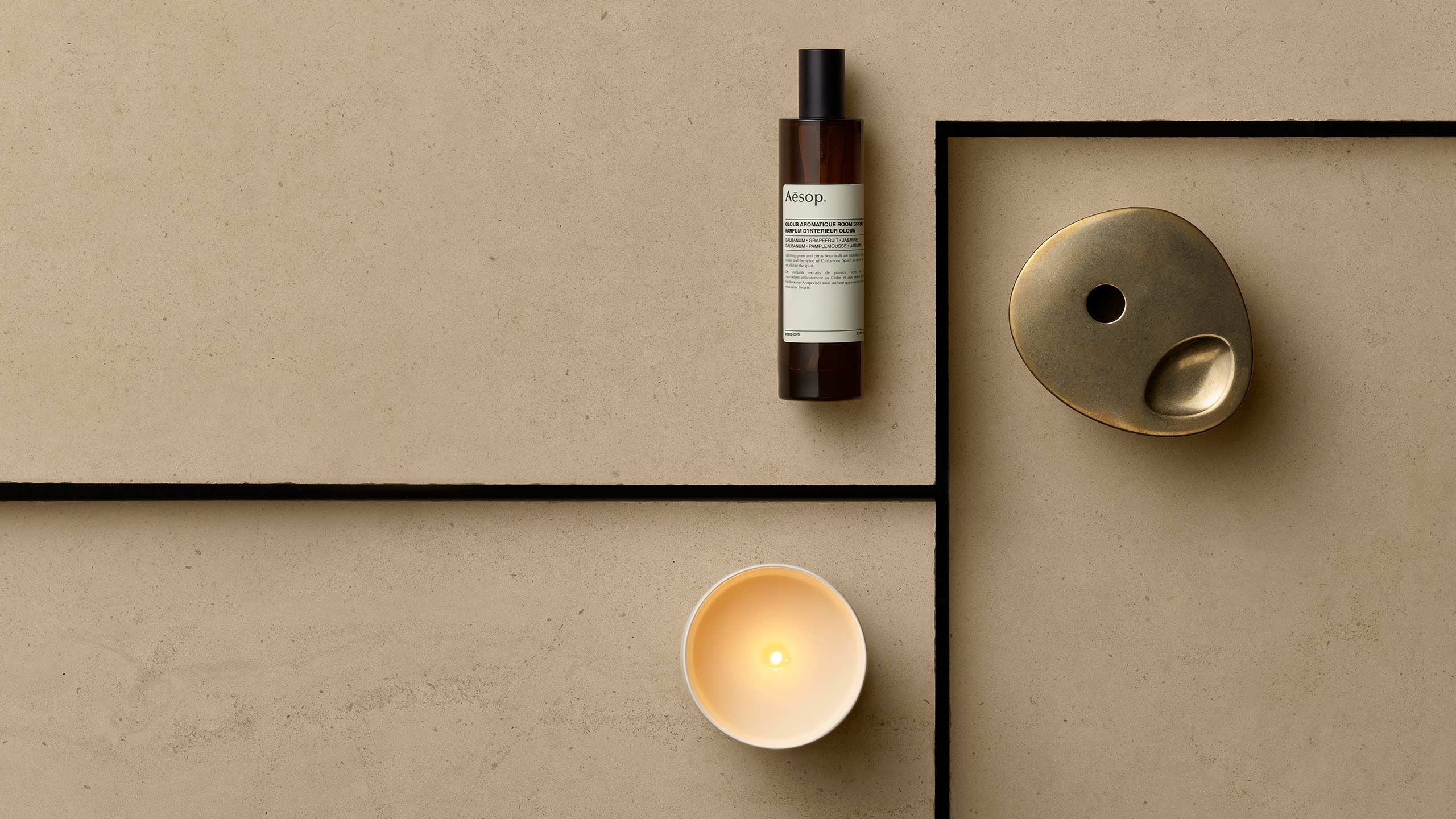 A lit Aromatique Candle sitting on a textured earth toned surface, alongside an amber bottle of Olous Aromatique Room Spray and Brass Oil Burner.