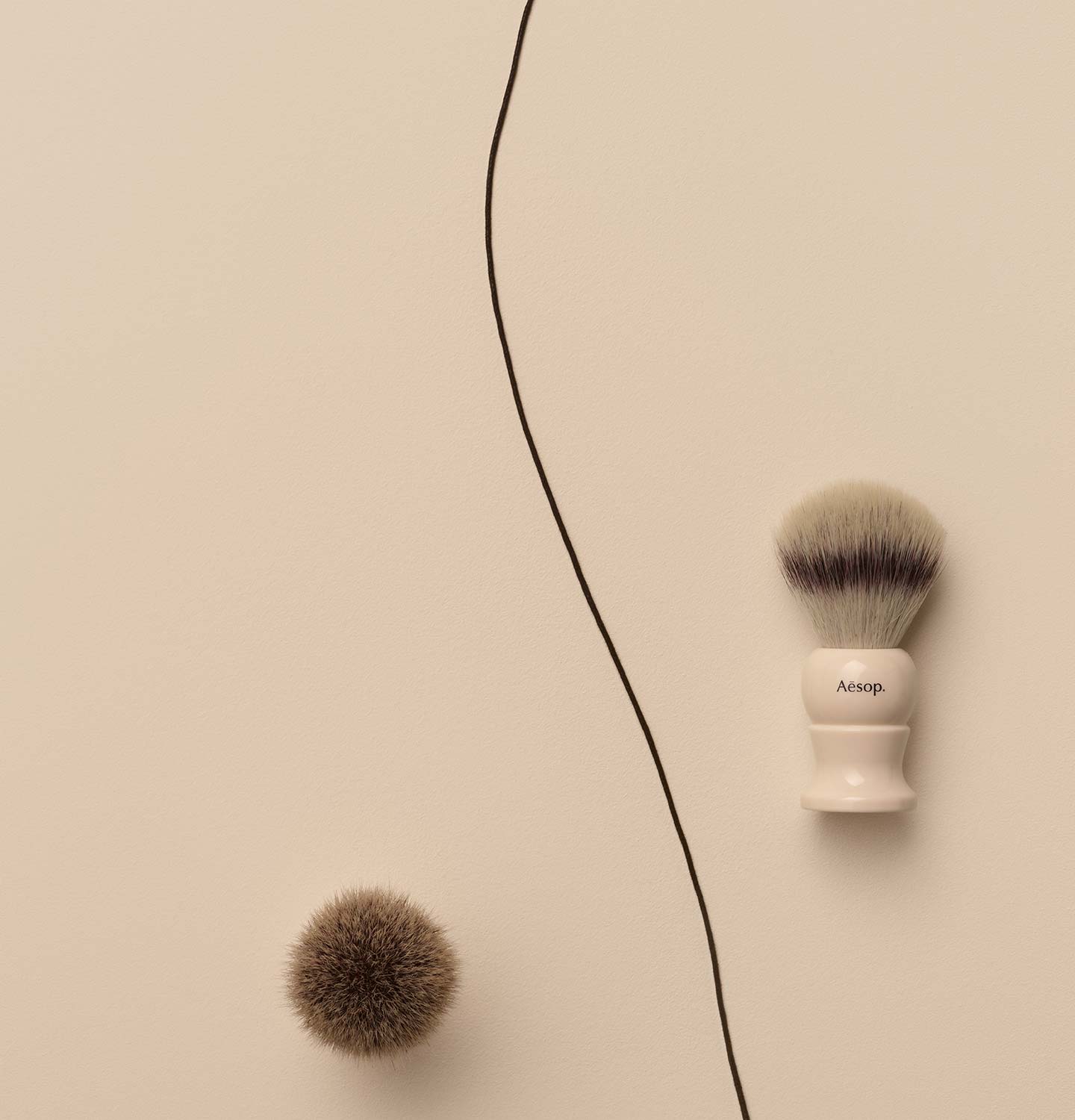 Aesop Shaving Brushes arranged alongside a dark string on a beige textured surface.