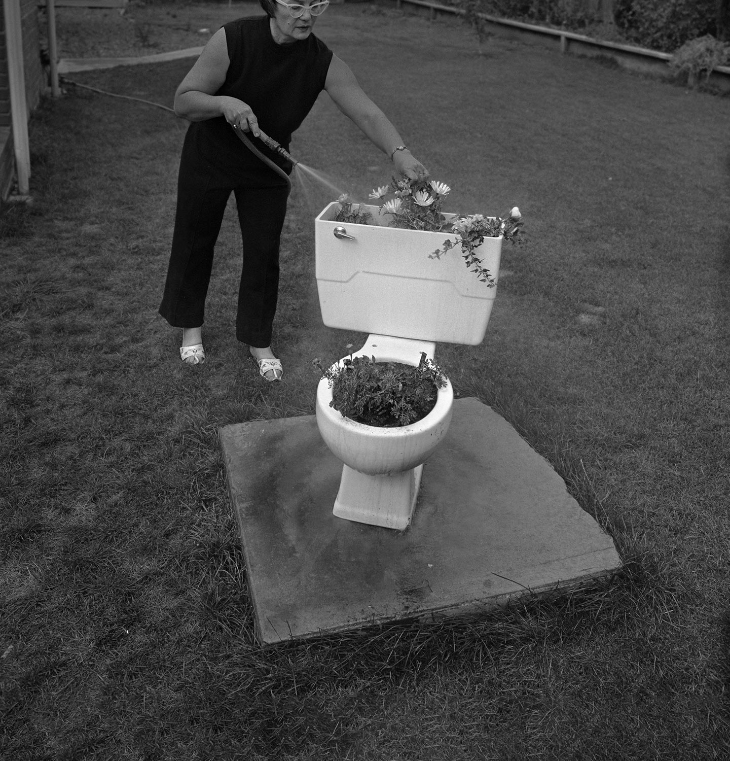 Retro black and white photograph depicting a woman outdoors using a hose to water flowers growing out of a toilet bowl.