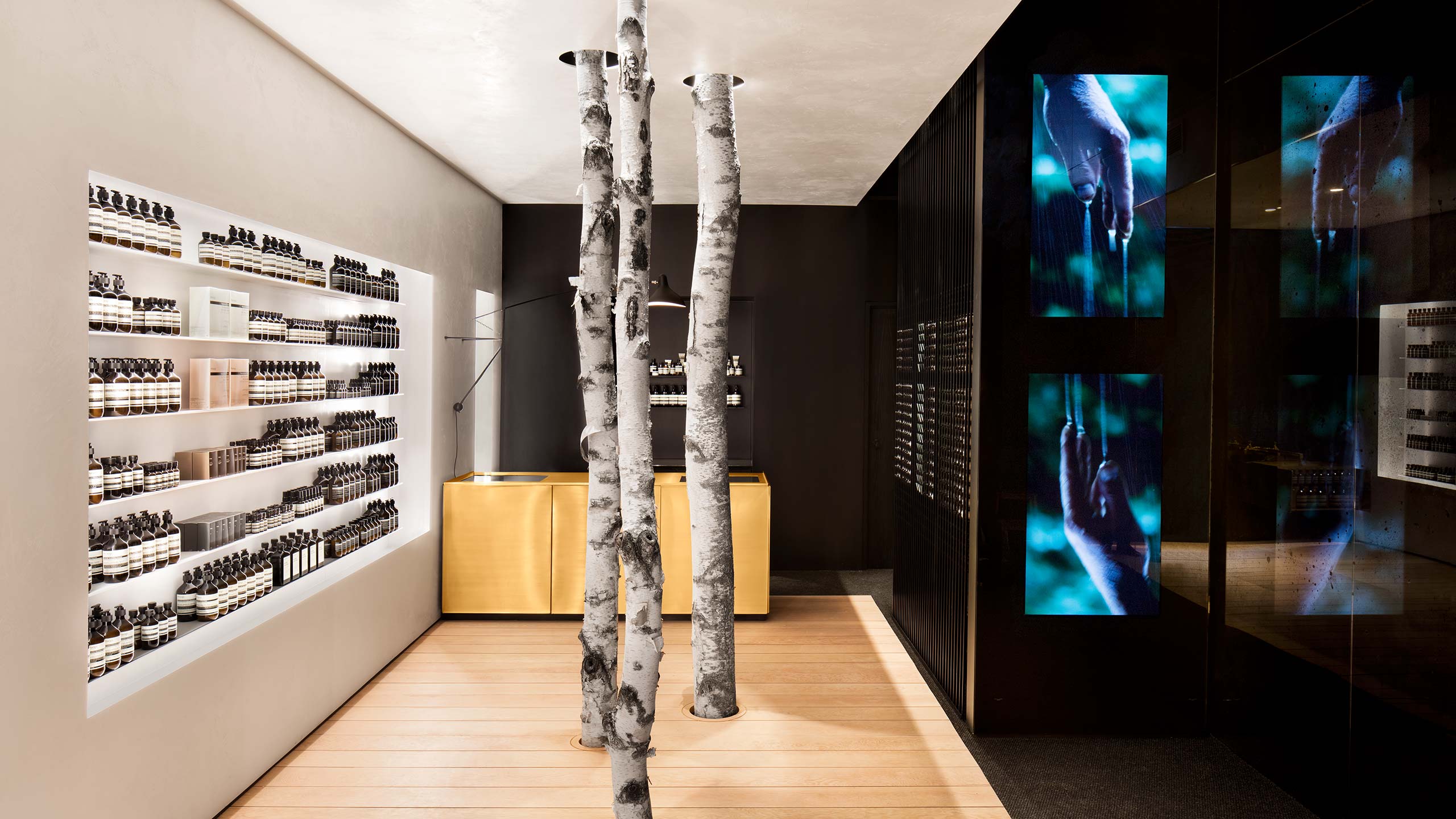 Three tall tree trunks acting as supporting beams within a modernist an Aesop store interior; contrasting with black mirror-like glass.