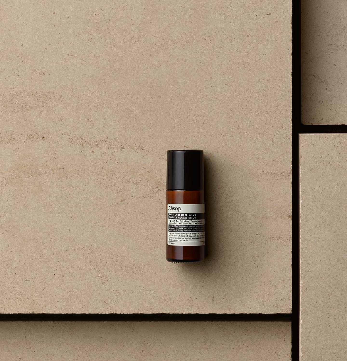 Aesop Herbal Deodorant Roll-On in amber bottle arranged on a beige textured surface.