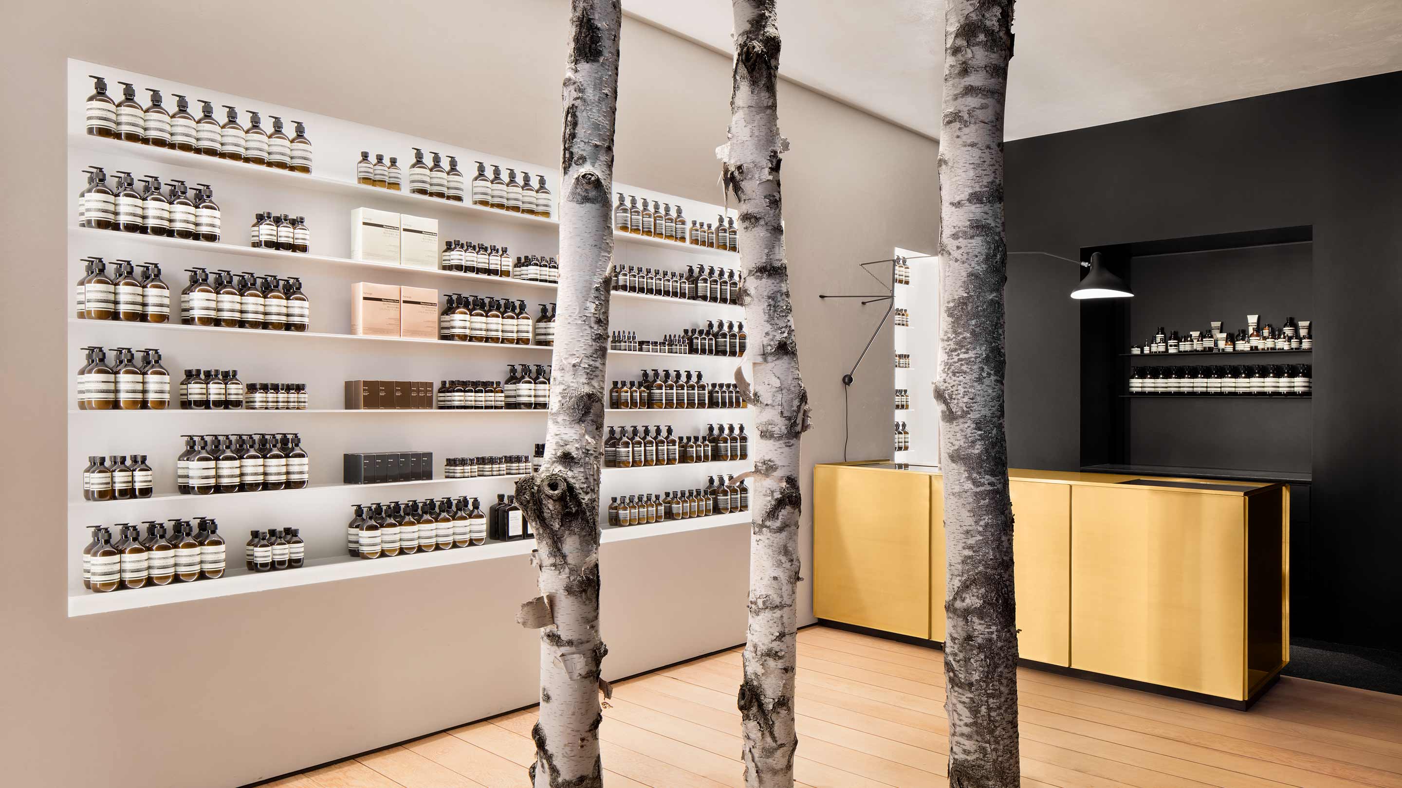 Three tall tree trunks acting as supporting beams within a modernist an Aesop store interior; fitted with brass counters and sinks against ice-black walls.