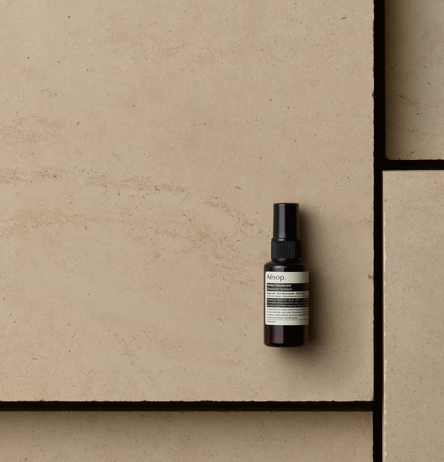 Aesop Herbal Deodorant in amber spray bottle arranged on a beige textured surface.