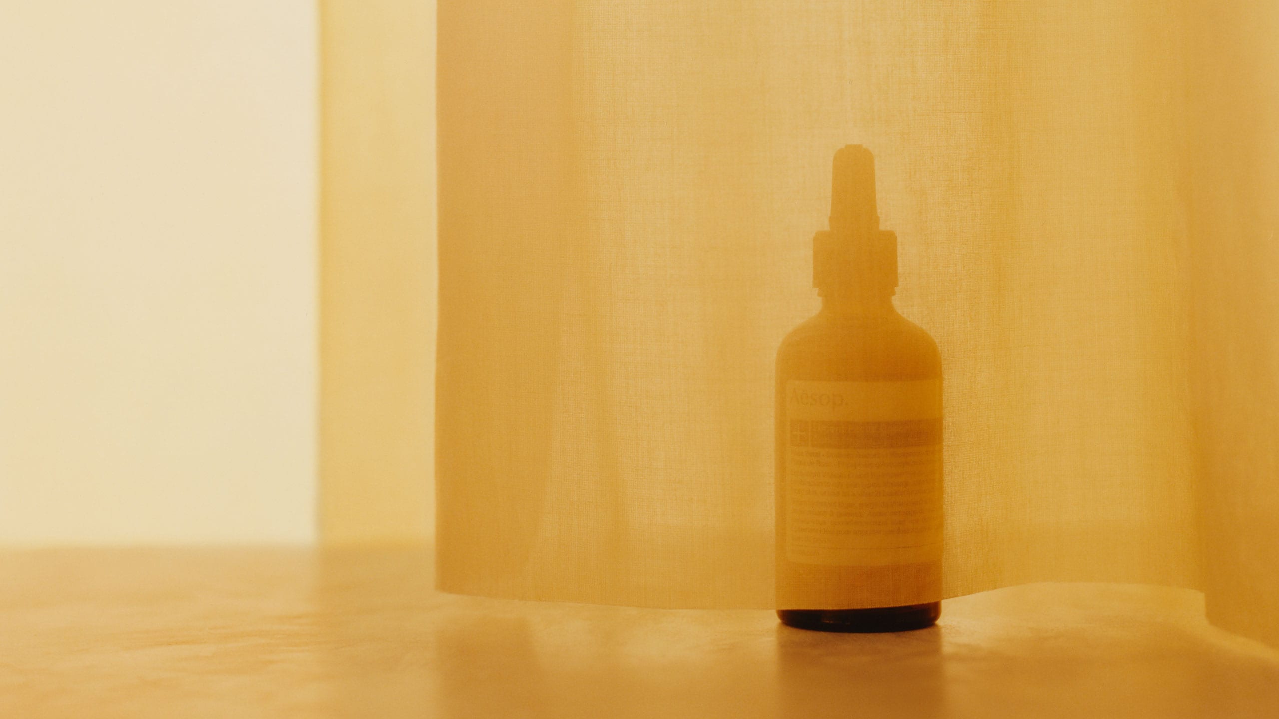 Aesop lucent facial concentrate in an amber glass bottle sitting behind a yellow curtain.