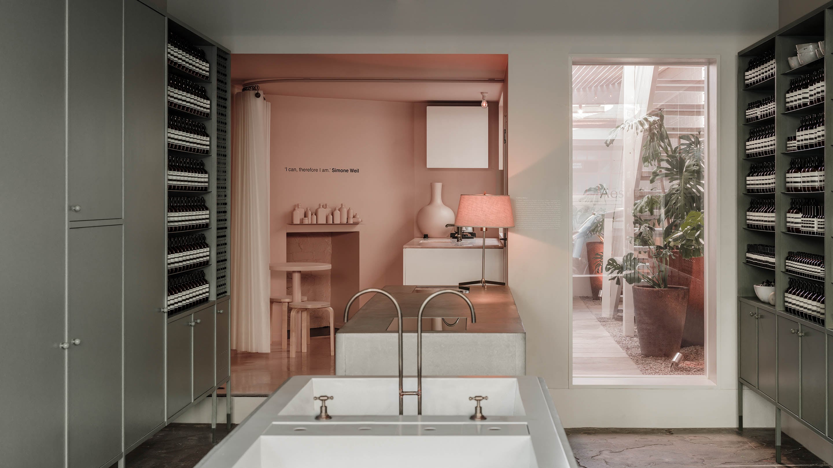 The cast concrete laundry-sink-styled basin sits in the middle of the store, flanked by pale green cupboards and shelves. In the background, a blush-pink kitchenette that features a silicon curtain.