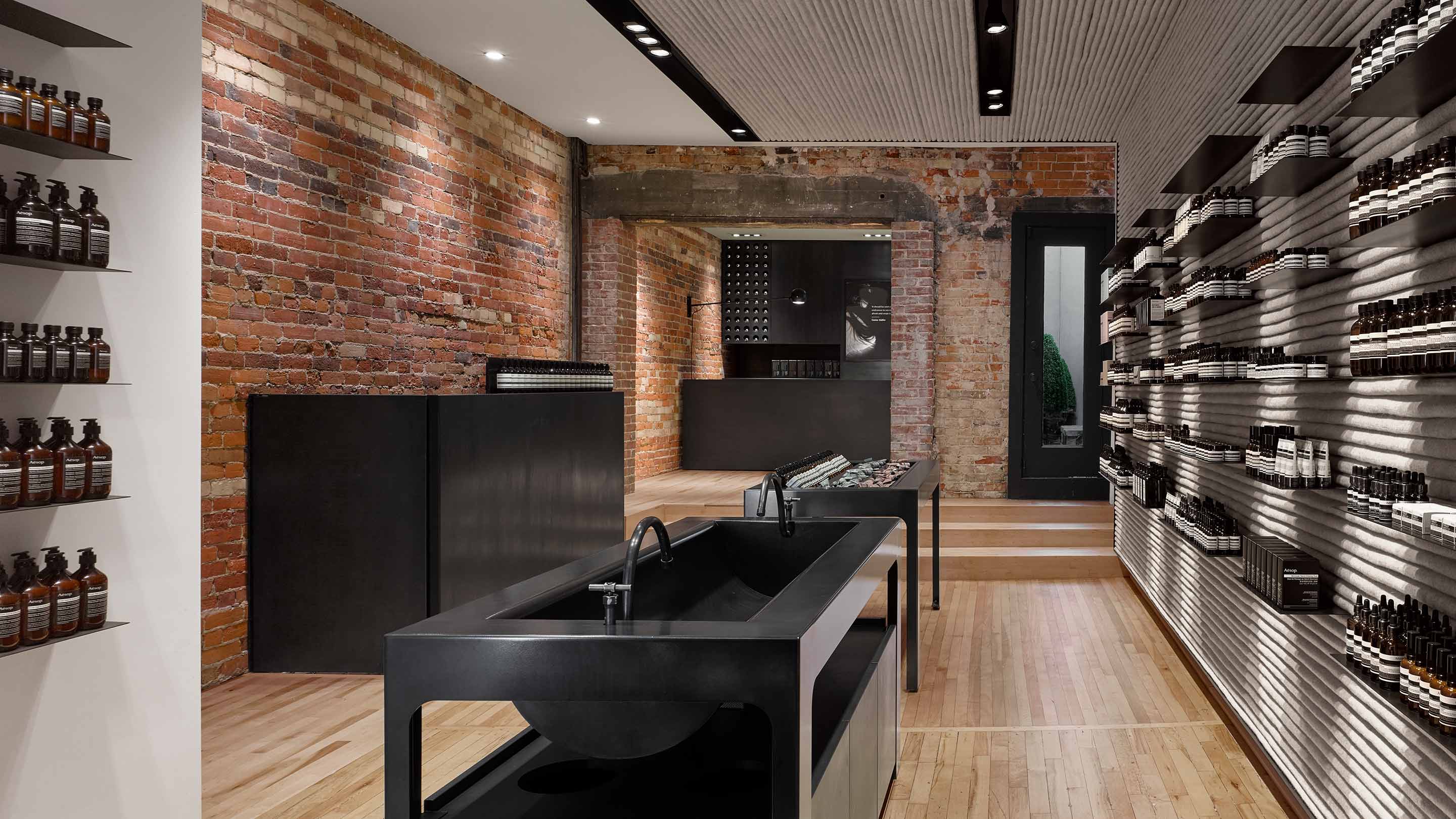 Smooth blackened steel sinks sit in the middle of a narrow store interior; contrasting with industrial textured brick walls and shelves holding Aesop Product.