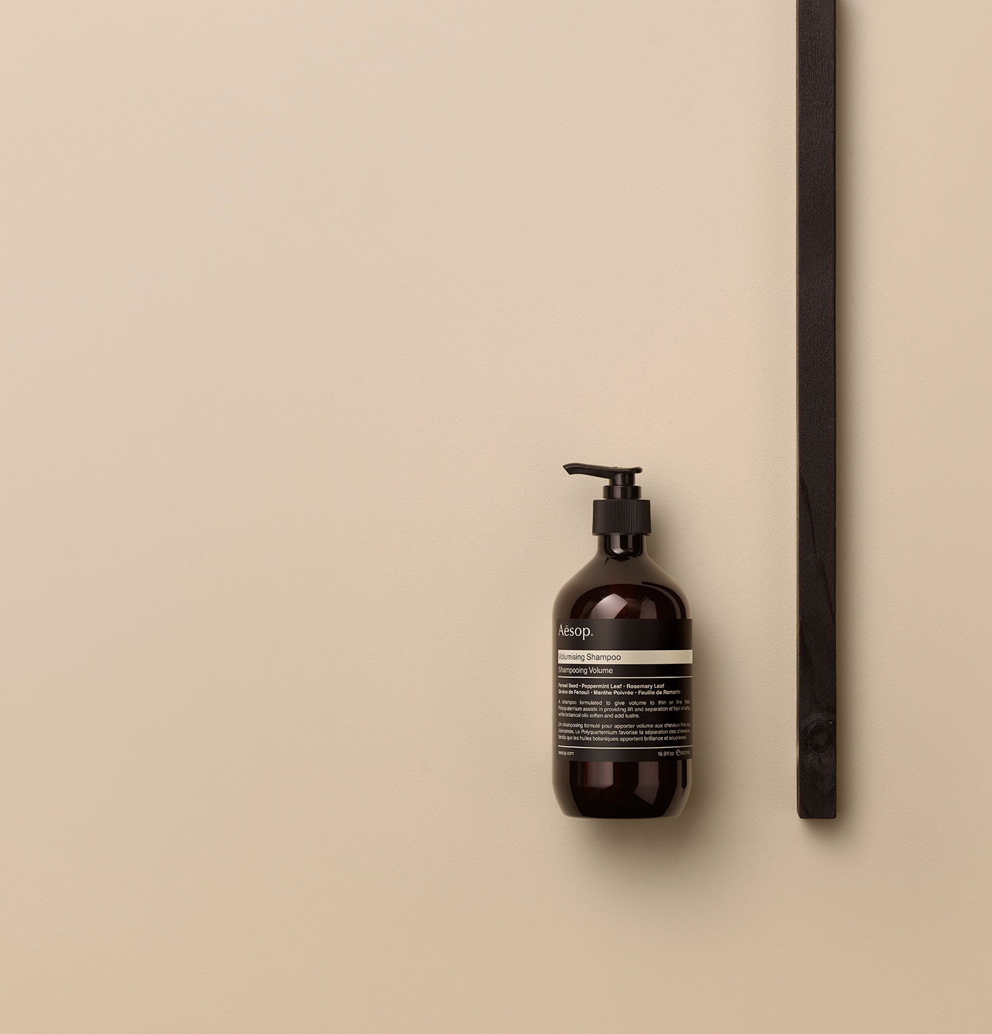 Aesop Volumising shampoo in amber pump bottle arranged alongside a dark wooden object on a beige surface.