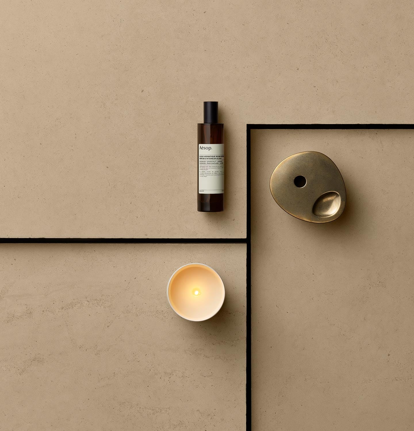 A lit Aromatique Candle sitting on a textured earth toned surface, alongside an amber bottle of Olous Aromatique Room Spray and Brass Oil Burner.