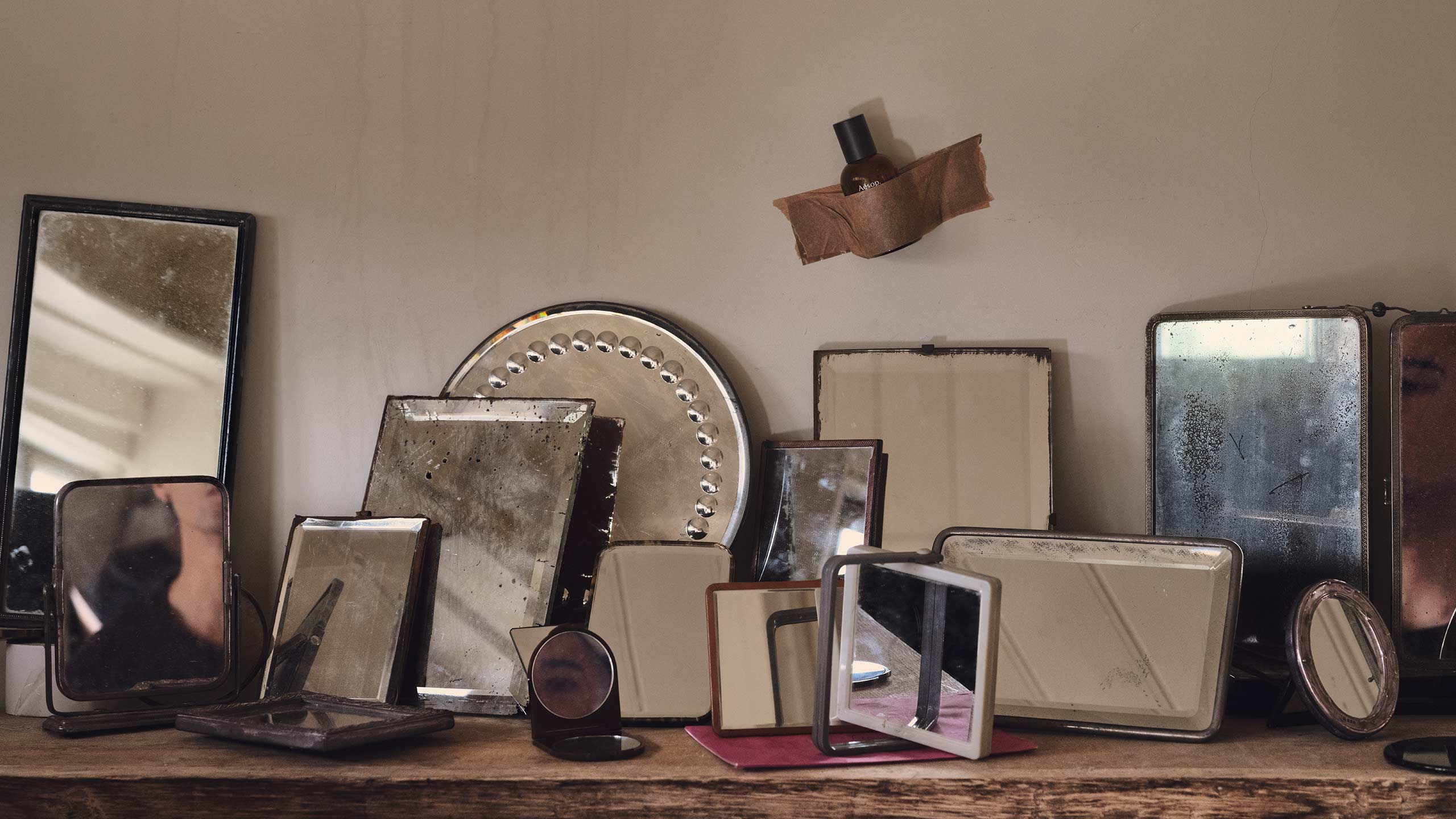 A glass bottle of Eidesis Eau de Parfum taped to the wall above an assortment of mirrors on a wooden bench.