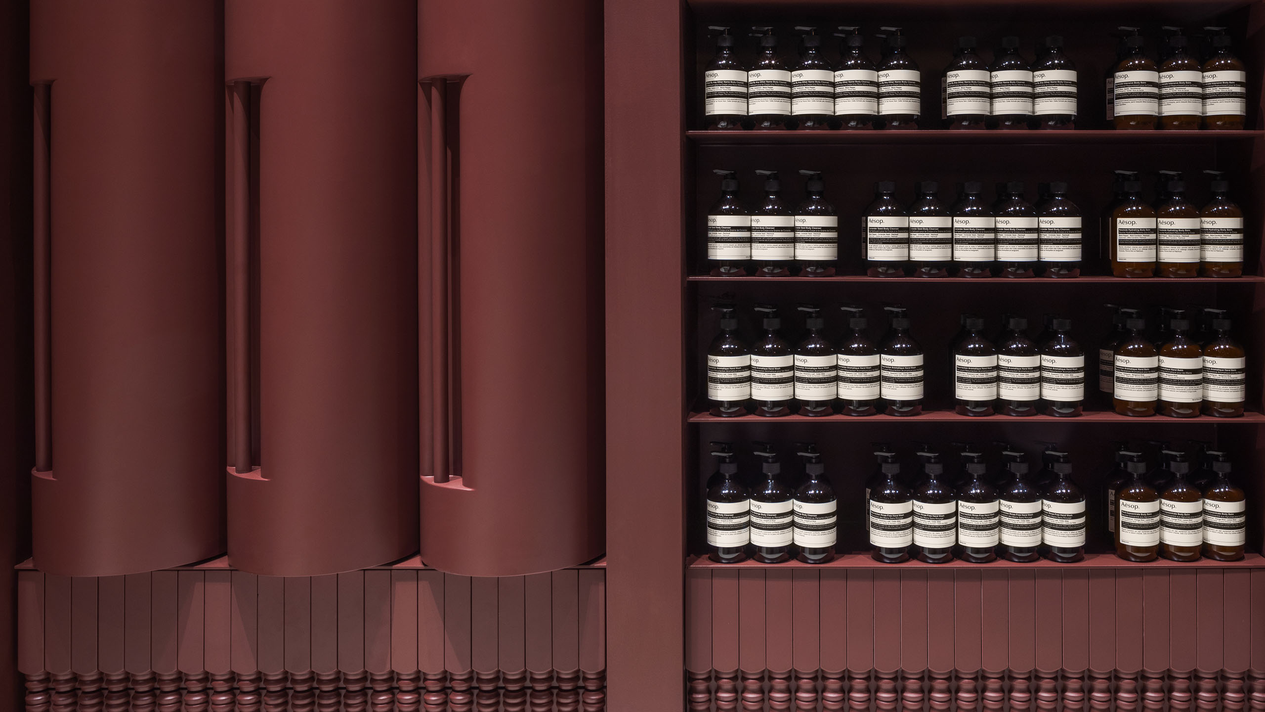 Three cylinder shelves are positioned on the left; rows of Aesop product are lined up on an open shelf to the right.