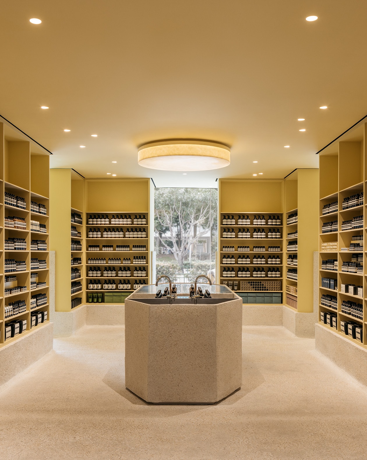 A large hand-cast pebble wash monolith in the middle of the Tampines store with two basins next to it and the smooth shelves on the surrounding walls are lined with Aesop products.