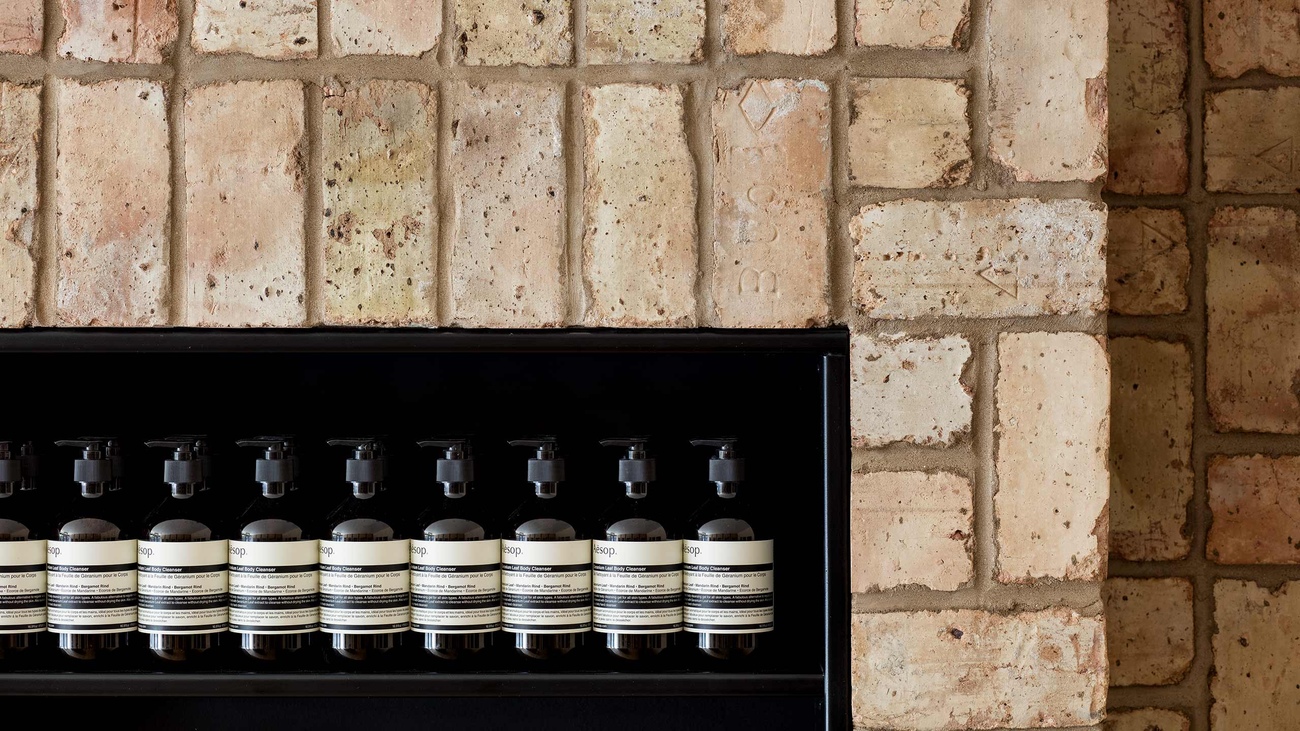 Nine pump bottles sit neatly aligned on a black steel shelf within a brick wall. 