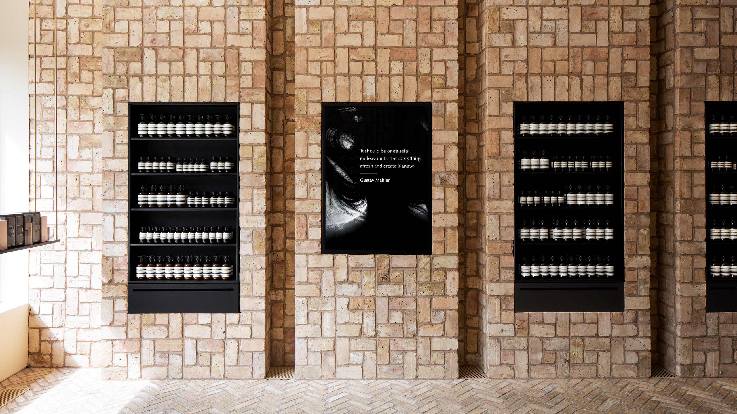 Two brick walls fitted with steel shelves holding Aesop product; middle brick wall fitted with light box. 