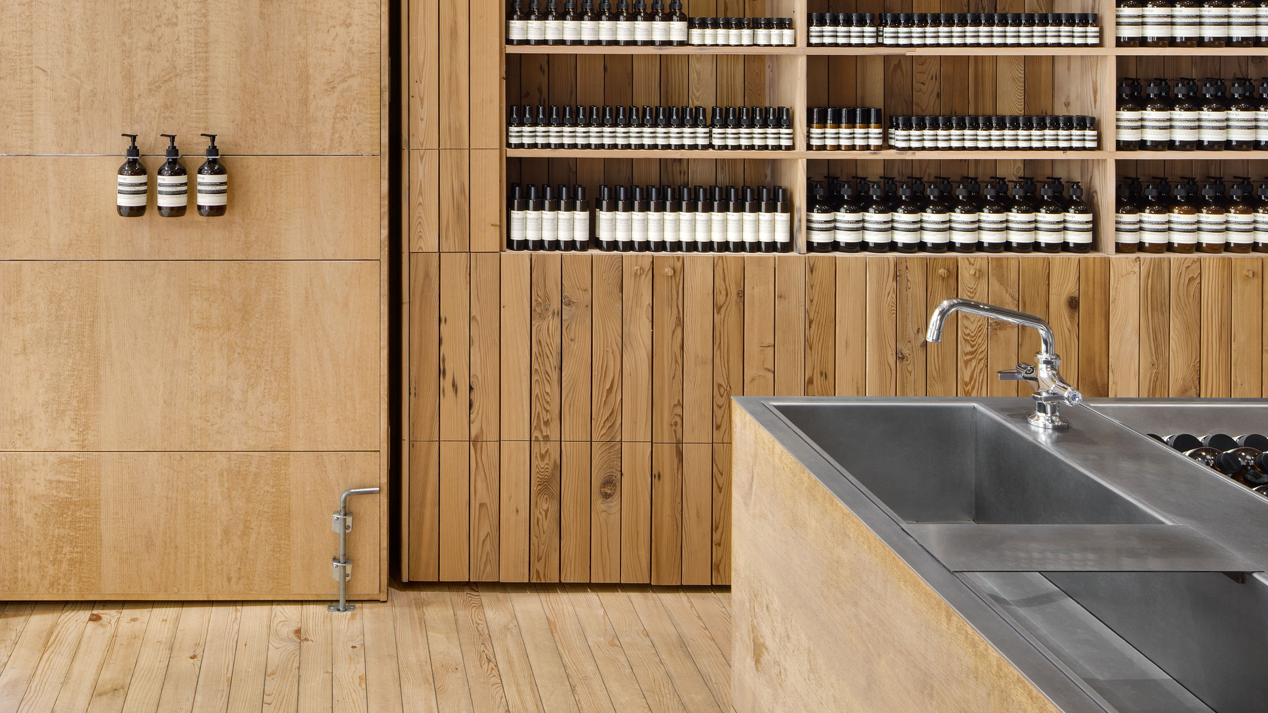 Three Aesop bottles sit together at the entrance door; steel sinks and shelves of Aesop product to the right