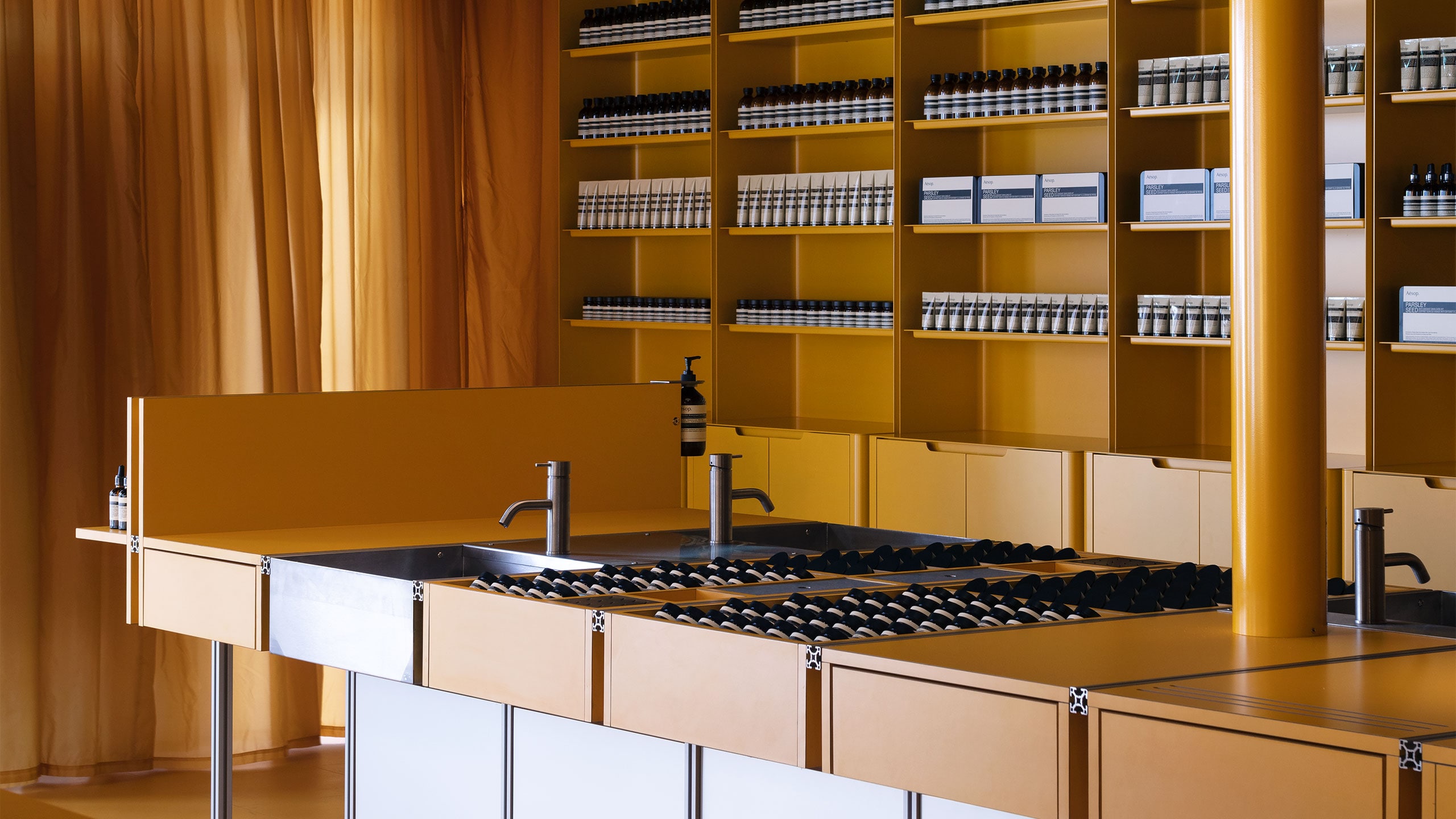 A powder-coated orange steel sink holds Aesop amber glass bottles with a large art-deco fibre glass light hangs above.