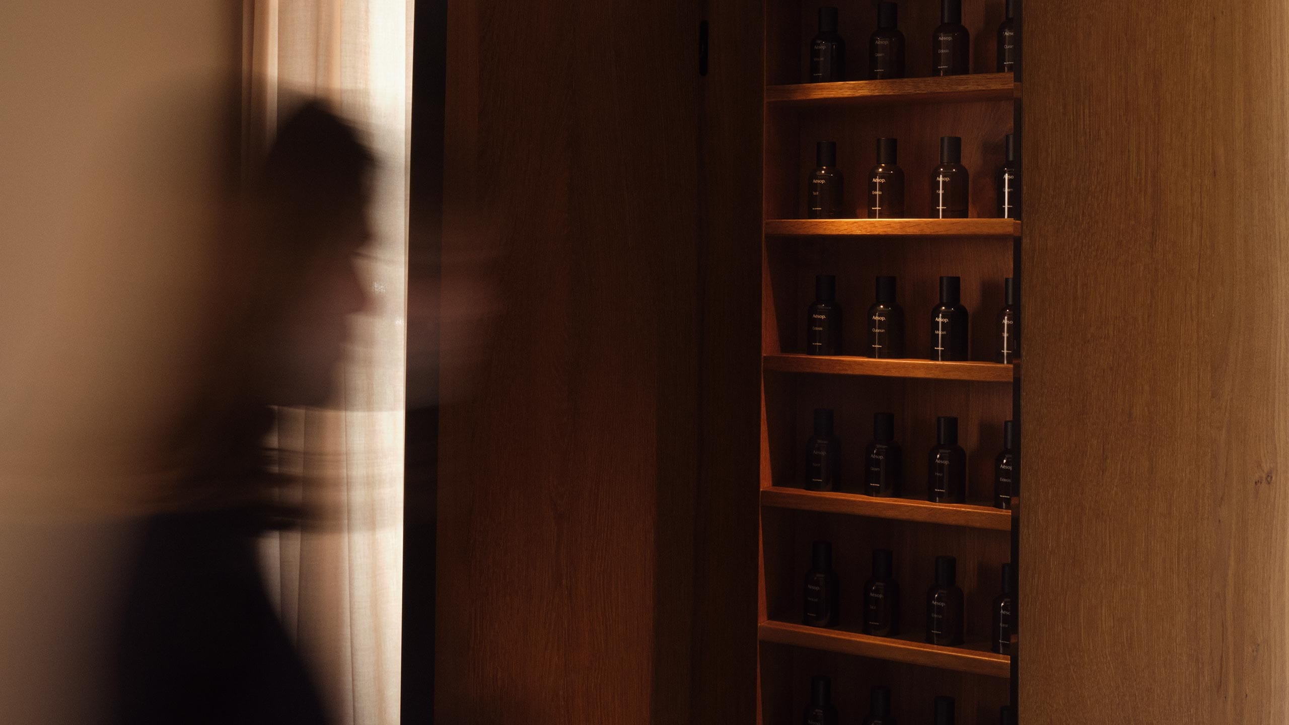 An individual walking past a wooden shelf displaying Aesop fragrances in amber bottles