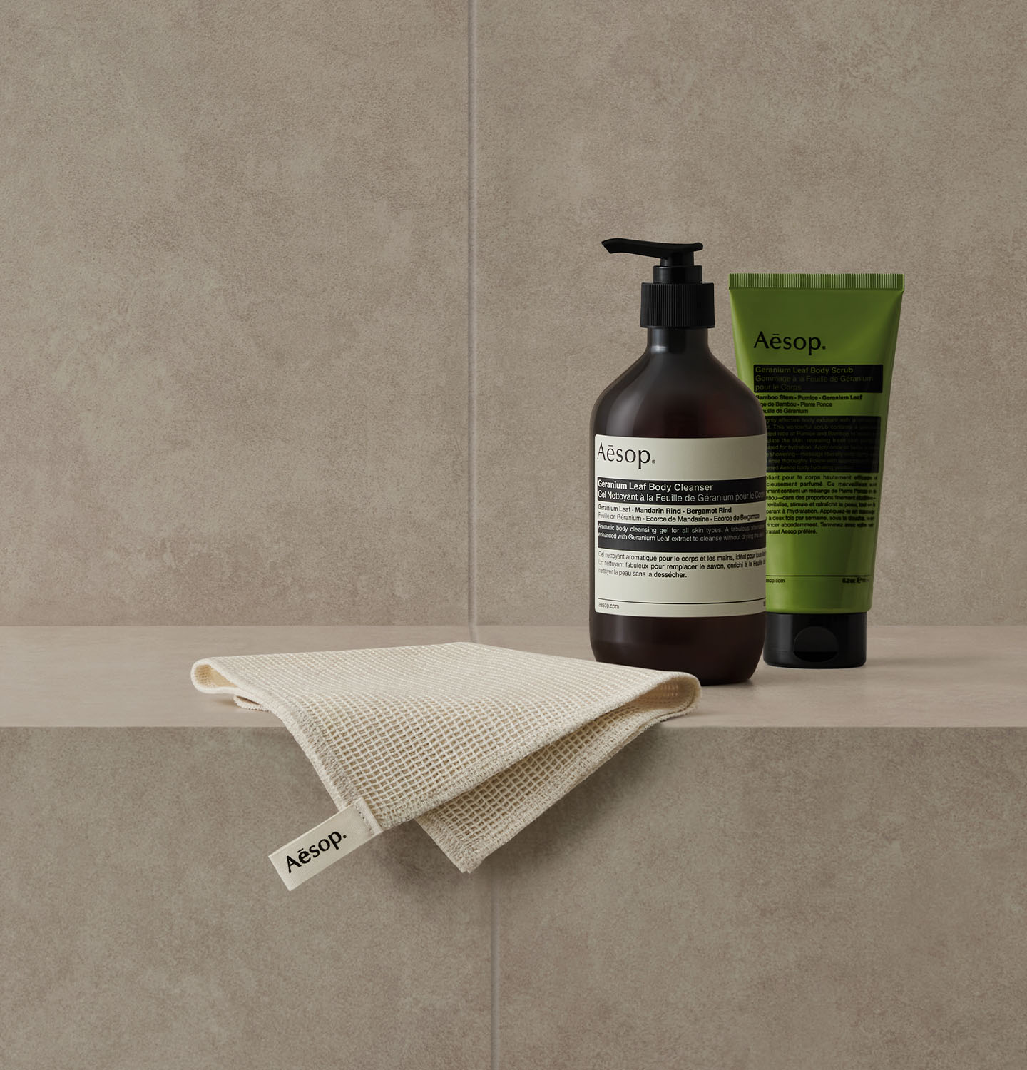 Geranium Leaf Body Cleanser and Geranium Leaf Body Scrub alongside Aesop shower towel placed on the grey bathroom tile. 