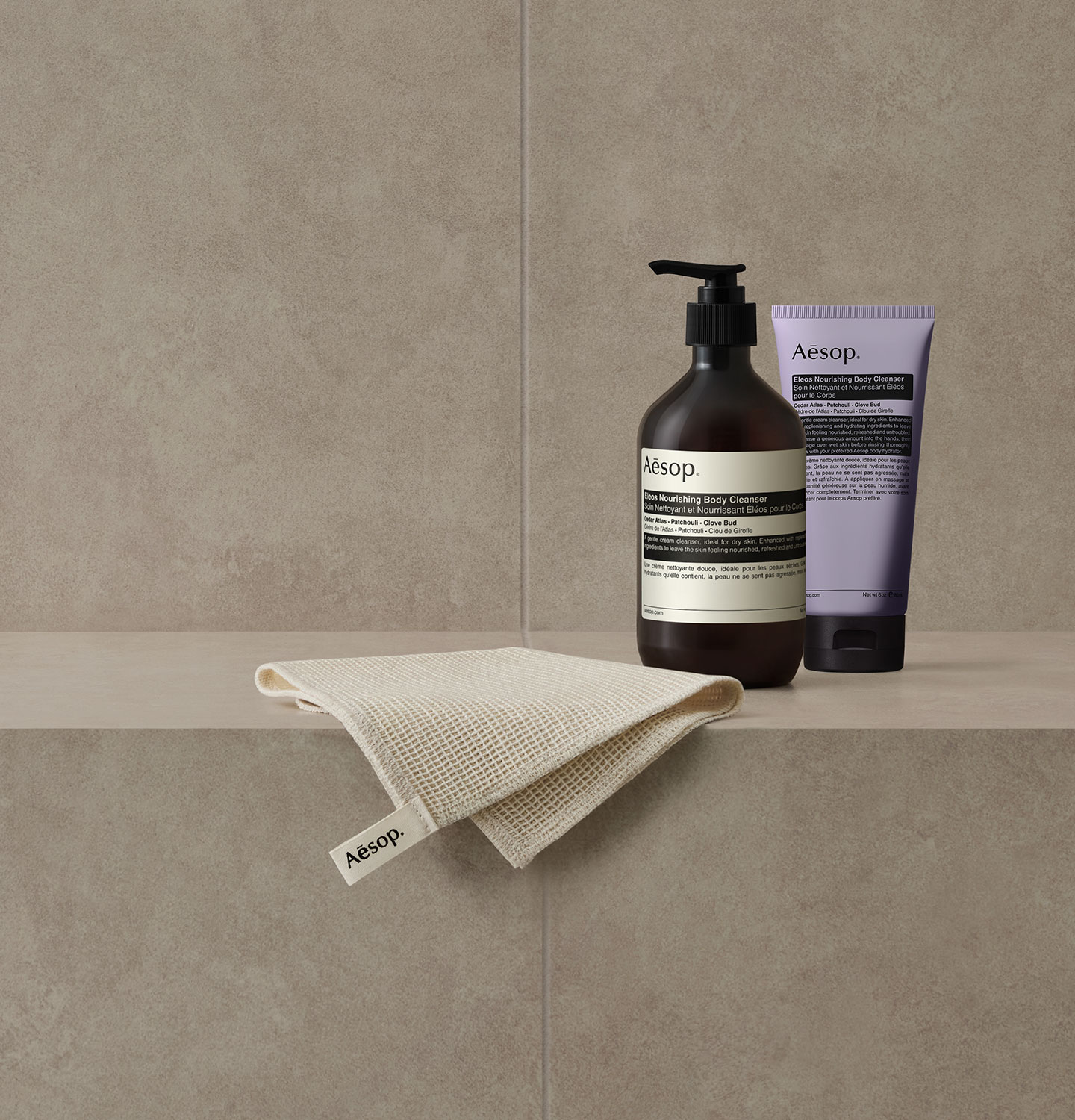 Eleos Nourishing Body Cleanser and Aesop shower towel being placed on the grey bathroom tile. 