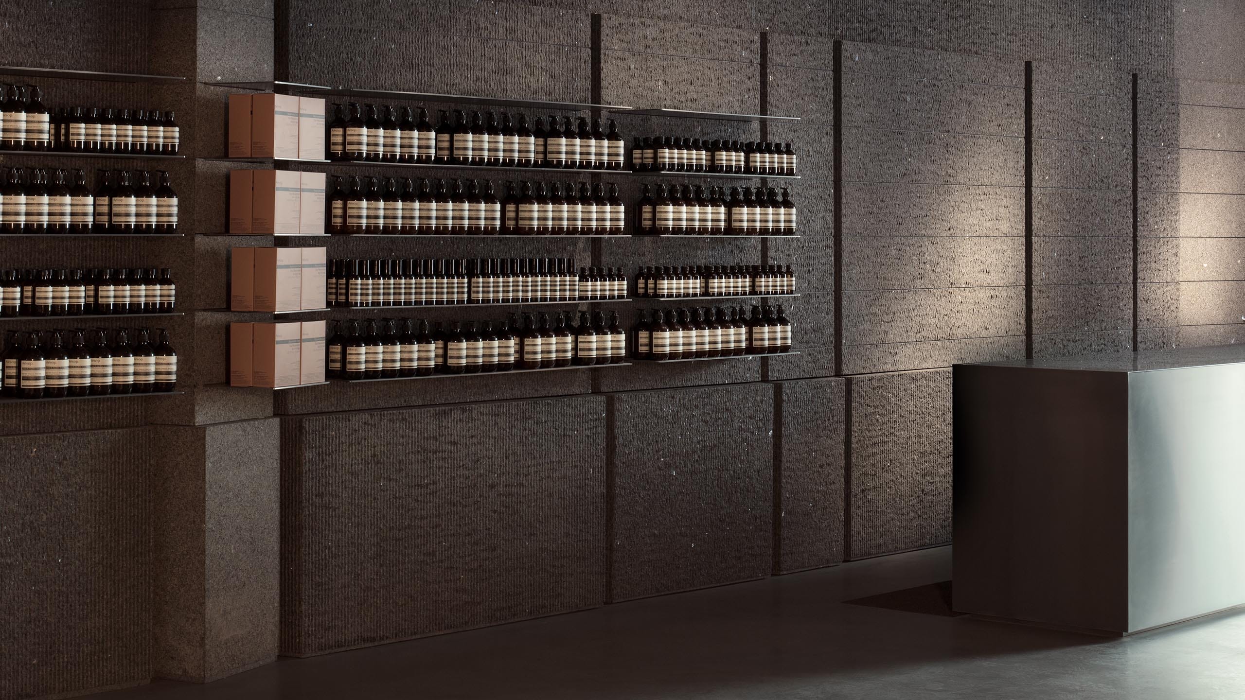 An ambient light shines on a steel sink and concrete shelving holding Aesop product