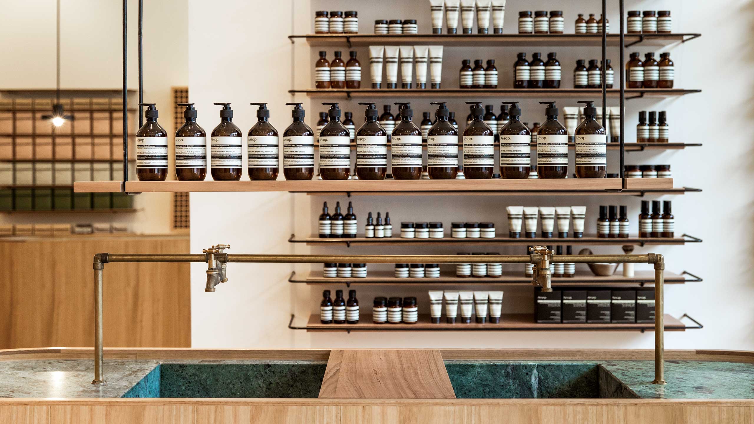 Several brown Aesop bottled on a shelf suspended by black metal poles. This shelf is suspended above marble basins.