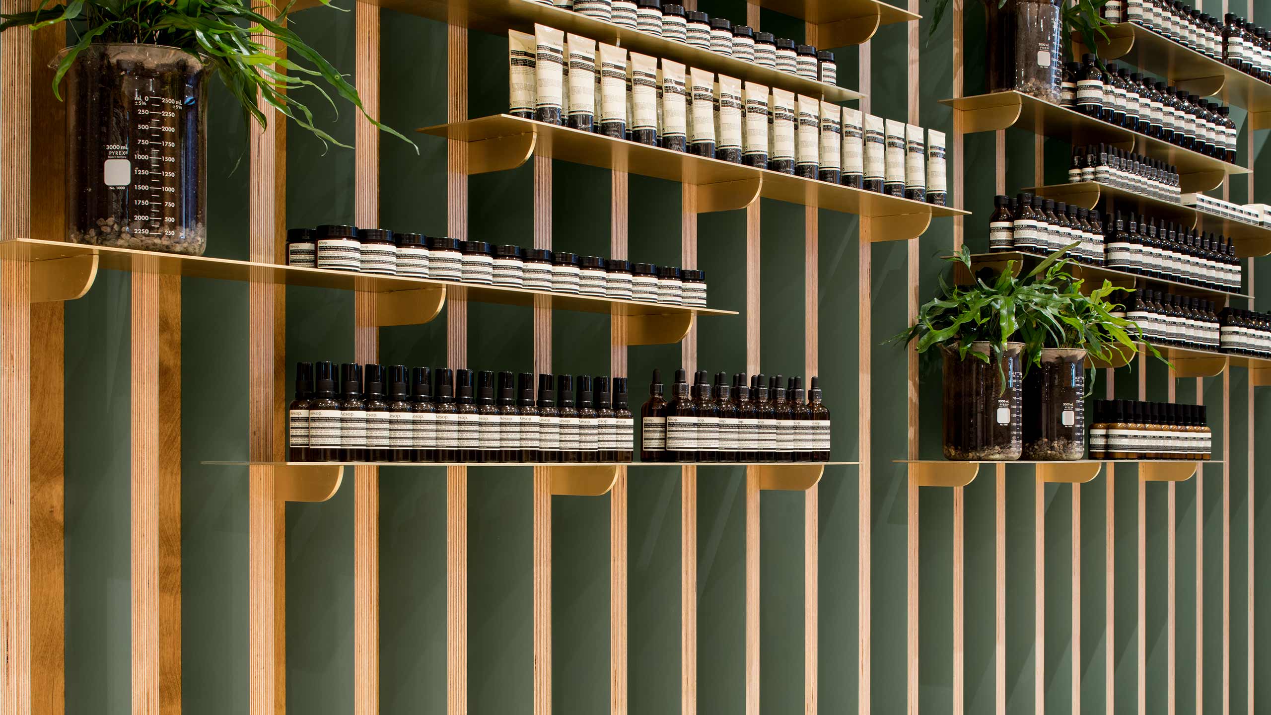 A stud wall made of plywood with contrasting subtle green panels; fitted with sheet metal shelving holding Aesop bottles.